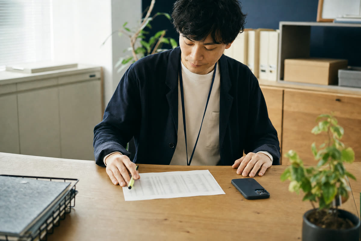 ぼかした名簿の紙の横に、伏せたスマートフォンと蛍光ペンが置かれた事務の机