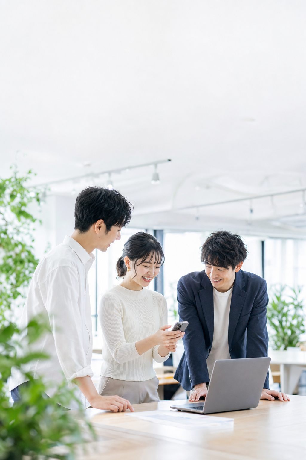 明るいオフィスで、スマホとパソコンを見ながら相談する3人（イメージ）
